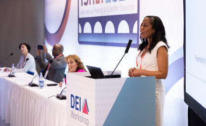 A black woman wearing a white dress stands behind a podium talking into a microphone