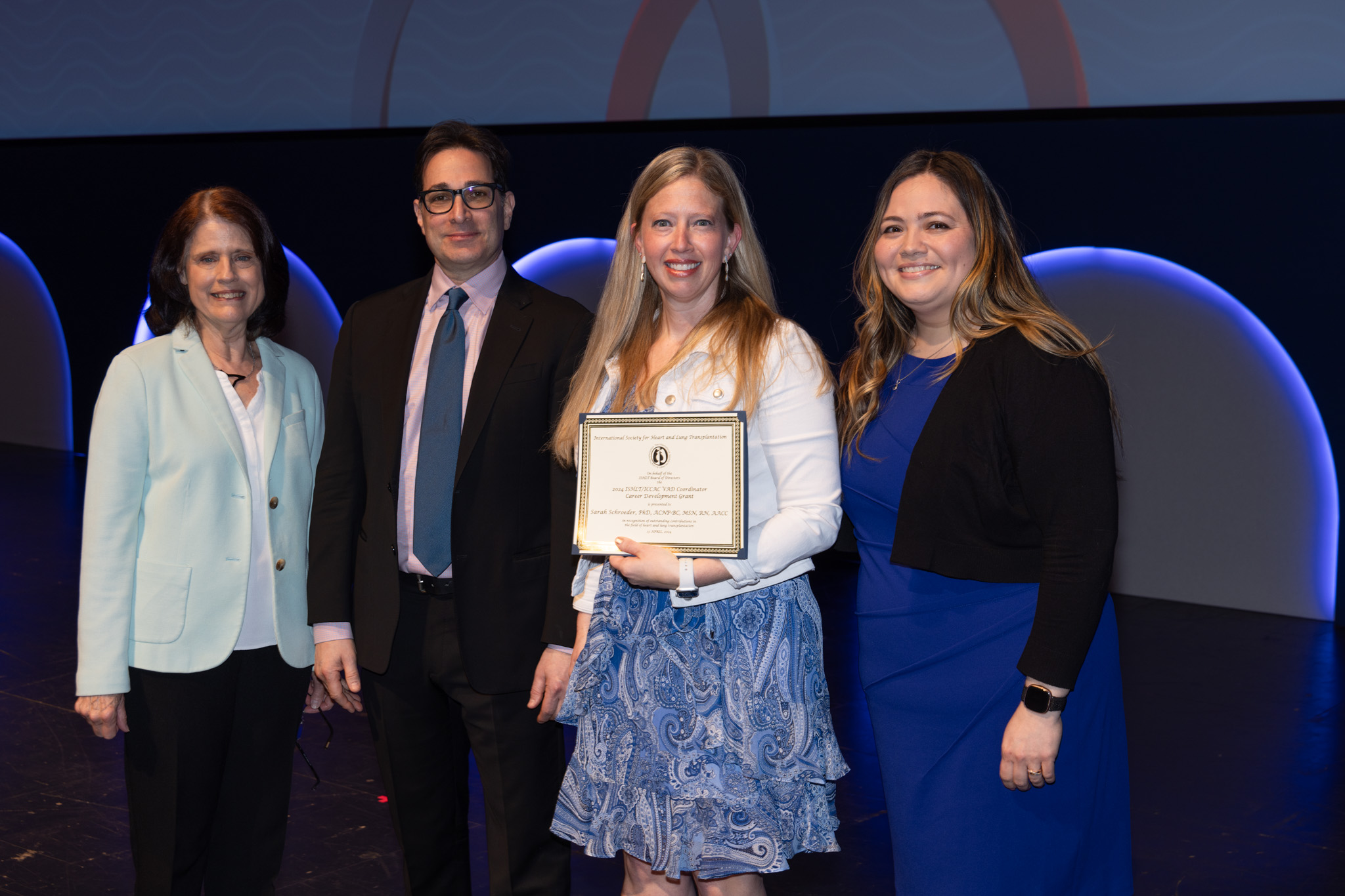 A woman with blonde hair is awarded a certificate at ISHLT2024 and poses with a woman with brown hair and a light blue suit jacket, a man with dark hair and a blue hair, and a woman with light brown hair and a blue dress