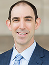 White man with brown hair posing in a light blue button down, stripped blue tie and navy blue sports coat