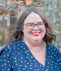 Woman with brown hair, wearing black glasses, red lipstick, and a blue polka dot blouse
