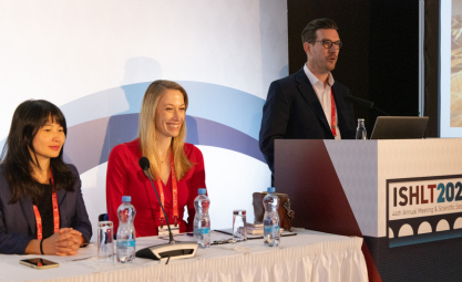 A white man with brown hair and black glasses speaks behind a podium while a blonde white woman and a red shirt and an Asian woman with dark hair sit beside him