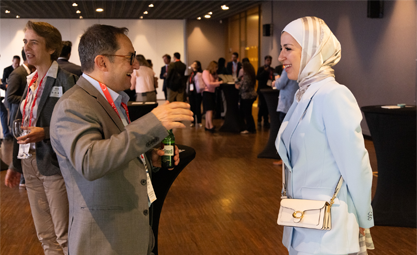 A man in a brown suit talks to a woman in a light blue suit and cream and gold scarf