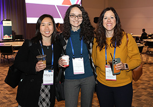 A young Asian woman in black long sleeve and patterned shirt, a white woman with curly hair wearing a blue shirt and grey pants, and aother white woman wearing a yellow long sleeve top and black pants pose together holding coffee cups